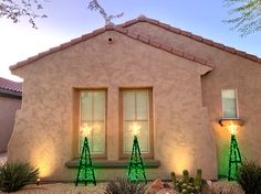 two green christmas trees sitting in front of a house with lights on it's windows