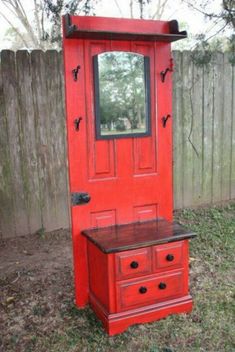 an old red cabinet with a mirror on the top and drawers below, in front of a wooden fence