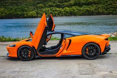 an orange sports car with its doors open sitting in front of a body of water