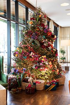 a christmas tree is decorated with colorful flowers and baskets on the floor in front of a large window