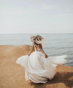 a woman in a white dress is walking on the sand by the ocean with her hair blowing in the wind