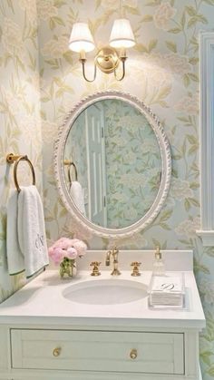 a bathroom with floral wallpaper and a round mirror above the sink, along with two gold faucets