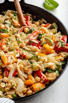 a skillet filled with pasta and vegetables on top of a white countertop next to a wooden spoon