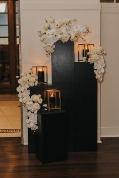 an arrangement of flowers and candles is displayed on a black pedestal in front of a white wall