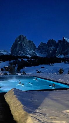 an outdoor swimming pool surrounded by snow covered mountains at night with lights on and people in the water