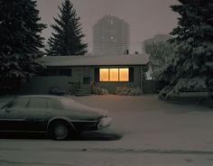 a car parked in front of a house on a snow covered street with trees and buildings behind it