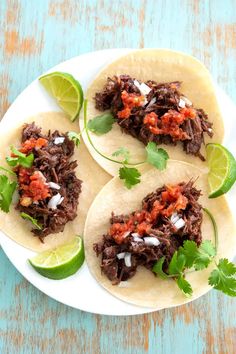 three shredded beef tacos on a white plate with limes and cilantro