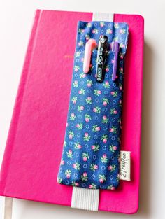 a pink and blue notebook with a pencil holder on it's cover, sitting on a white surface