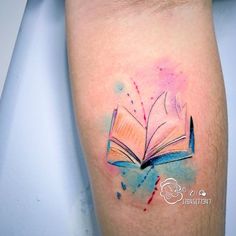 an open book tattoo on the left arm with watercolor paint splatters around it