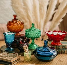 there are many different colored glass bowls on the table next to some books and feathers