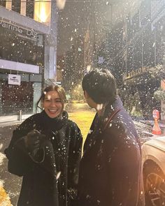 two people standing in the snow talking to each other on a city street at night