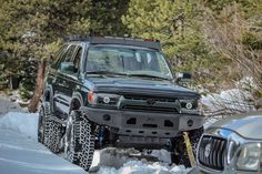 an suv is parked in the snow next to some trees and bushes, while another vehicle sits behind it