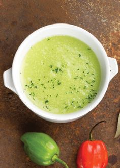 a white bowl filled with green soup next to two red peppers on top of a table