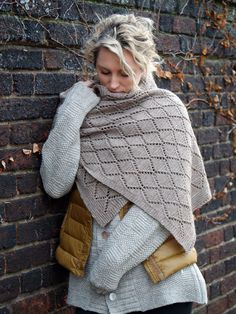 a woman leaning against a brick wall wearing a knitted shawl and vest with pockets