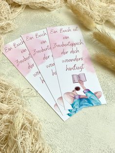 three pink and white tags with german words on them sitting next to some brown feathers