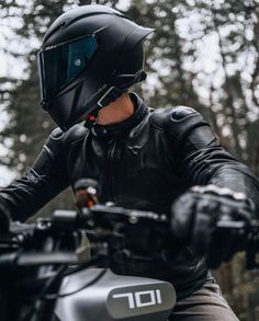 a man wearing a black leather jacket and helmet sitting on a motorbike with trees in the background