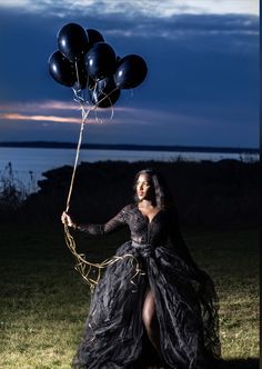 a woman in a long black dress holding balloons and posing for the camera at night