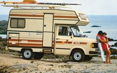 a man and woman standing next to an rv parked on top of a rocky beach