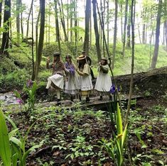 three people dressed in period costumes walking across a bridge over a stream through the woods