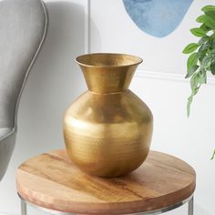 a gold vase sitting on top of a wooden table next to a gray chair and potted plant