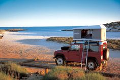 a red truck parked on top of a sandy beach next to the ocean with a white roof