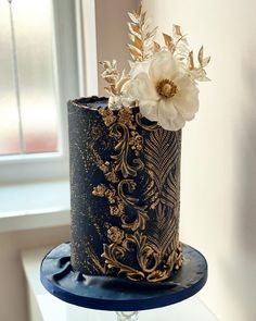 a black and gold wedding cake with white flowers on the top is sitting in front of a window