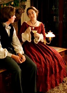 a man and woman dressed in period clothing sitting on a bench talking to each other