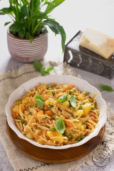 a white bowl filled with pasta and vegetables on top of a wooden table next to a potted plant