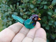 a small glass bird sitting on top of someone's hand in front of some bushes