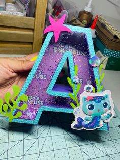 a hand holding up a purple and blue letter shaped like a mermaid with sea life around it