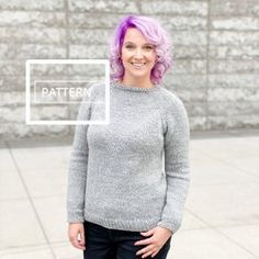 a woman with purple hair standing in front of a brick wall wearing a gray sweater and black pants