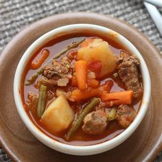 a bowl of stew with meat, potatoes and green beans on a plate next to a fork