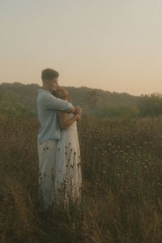 a man and woman standing in the middle of a field at sunset, hugging each other