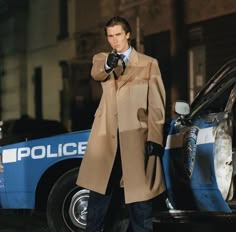 a man in a trench coat standing next to a police car and pointing at the camera