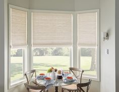 a dining room table set for four in front of three large windows with roman shades