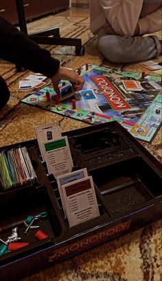 two people sitting on the floor playing monopoly with their hands in an open box that is filled with cards and other items