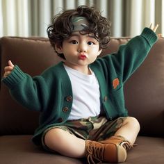 a little boy sitting on top of a couch wearing a green cardigan sweater and camo shorts