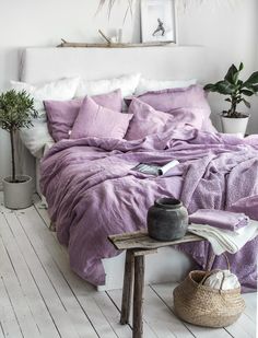 a bed with purple sheets and pillows in a white room next to a potted plant