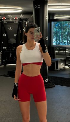 a woman taking a selfie with her cell phone in a gym room while wearing red and white shorts