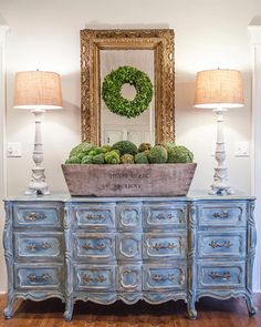 a blue dresser with a wreath on top and two lamps next to it in front of a mirror