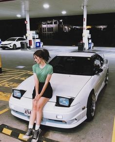 a woman sitting on the hood of a white car in a gas station parking lot