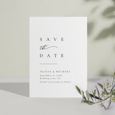 a white save the date card with greenery on it next to an olive branch