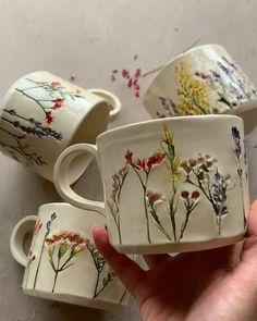 three hand painted coffee cups and saucers with wildflowers on them are being held by a person