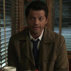 a man in a trench coat and tie sitting at a table with a coffee cup