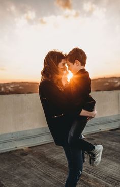 a man holding a woman in his arms as the sun sets behind them on top of a roof