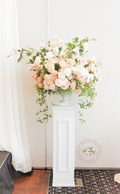 a white pedestal with flowers in it sitting on a rug next to a wall and window