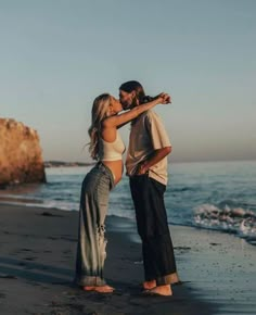 a man and woman standing on top of a sandy beach next to the ocean with their arms around each other