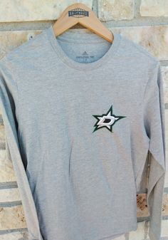 Show off your team pride in this Dallas Stars Grey Jersey History Long Sleeve T Shirt! This Dallas Stars Long Sleeve Tee features a lightweight, cotton material with a crew neckline, long sleeves, and screen printed team graphics. Make sure everyone knows you root for the Stars with this Grey Dallas Stars T Shirt. Go Stars!! Soft hand, ring spun cotton material, Regular fit, Crew neckline, Rib-knit collar, Screen print of team logo on left chest, Screen print of team graphic down right sleeve, M Adidas Cotton T-shirt For Fan Merchandise, Adidas Cotton Tops For Sports Season, Adidas Crew Neck Top, Athletic Heather Long Sleeve Cotton T-shirt, Casual Adidas Tops For Fan Merchandise, Casual Adidas Tops For Fans, Grey Cotton Long Sleeve T-shirt, Adidas Long Sleeve Cotton T-shirt, Adidas Gray Cotton Tops