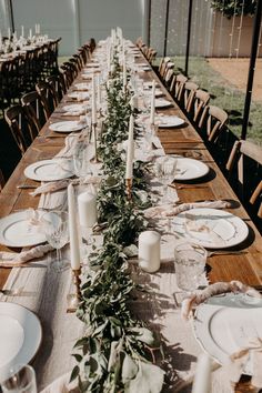 a long wooden table with white plates and greenery on it, surrounded by candles