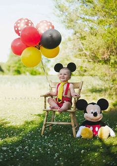 a baby sitting in a chair with mickey mouse and balloons on the grass near him